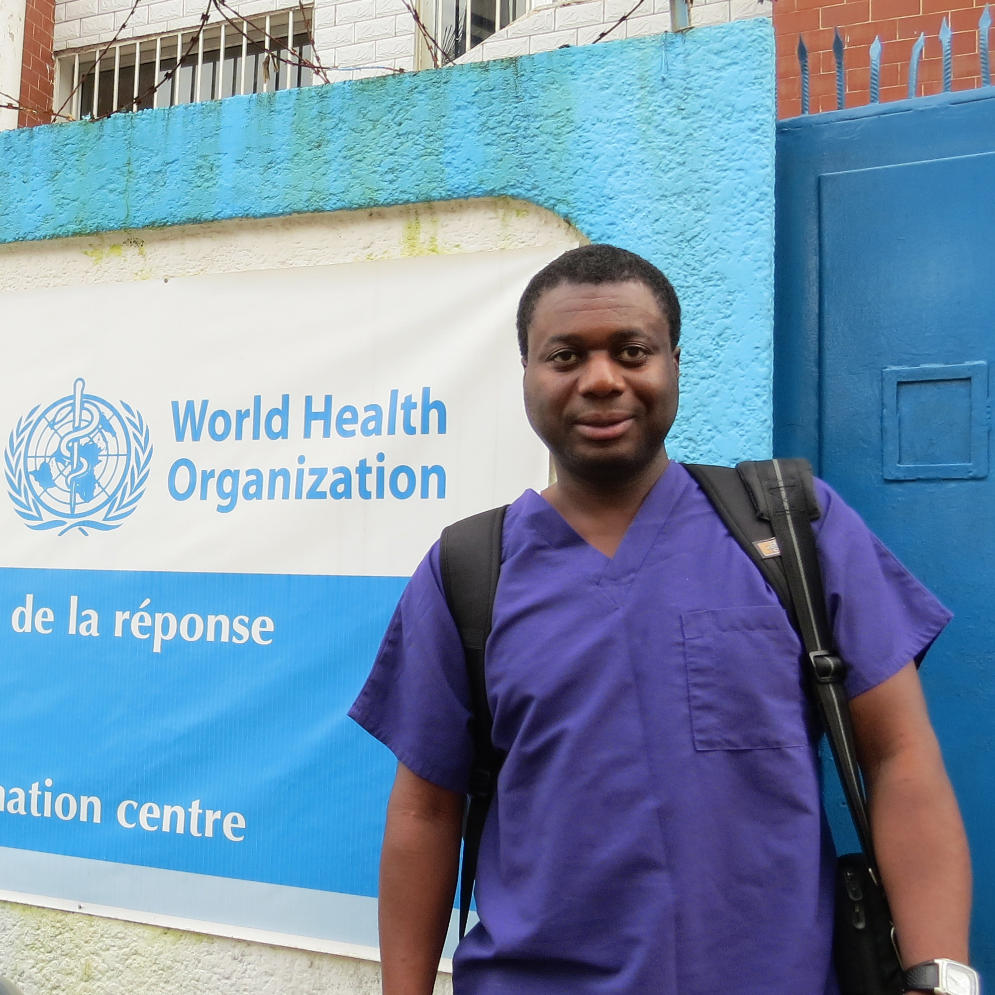 Man wearing medical scrubs stands in front of a World Health Organization poster