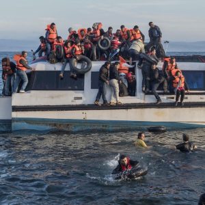 Dozens of refugees cling to sides and top of an old motorboat in the Mediterranean Sea. Several people are in the water beside the boat.