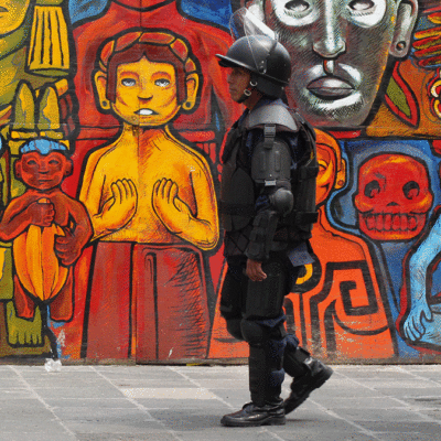 Two police officers in riot gear standing in front of a mural of people in an indigenous Mexican style