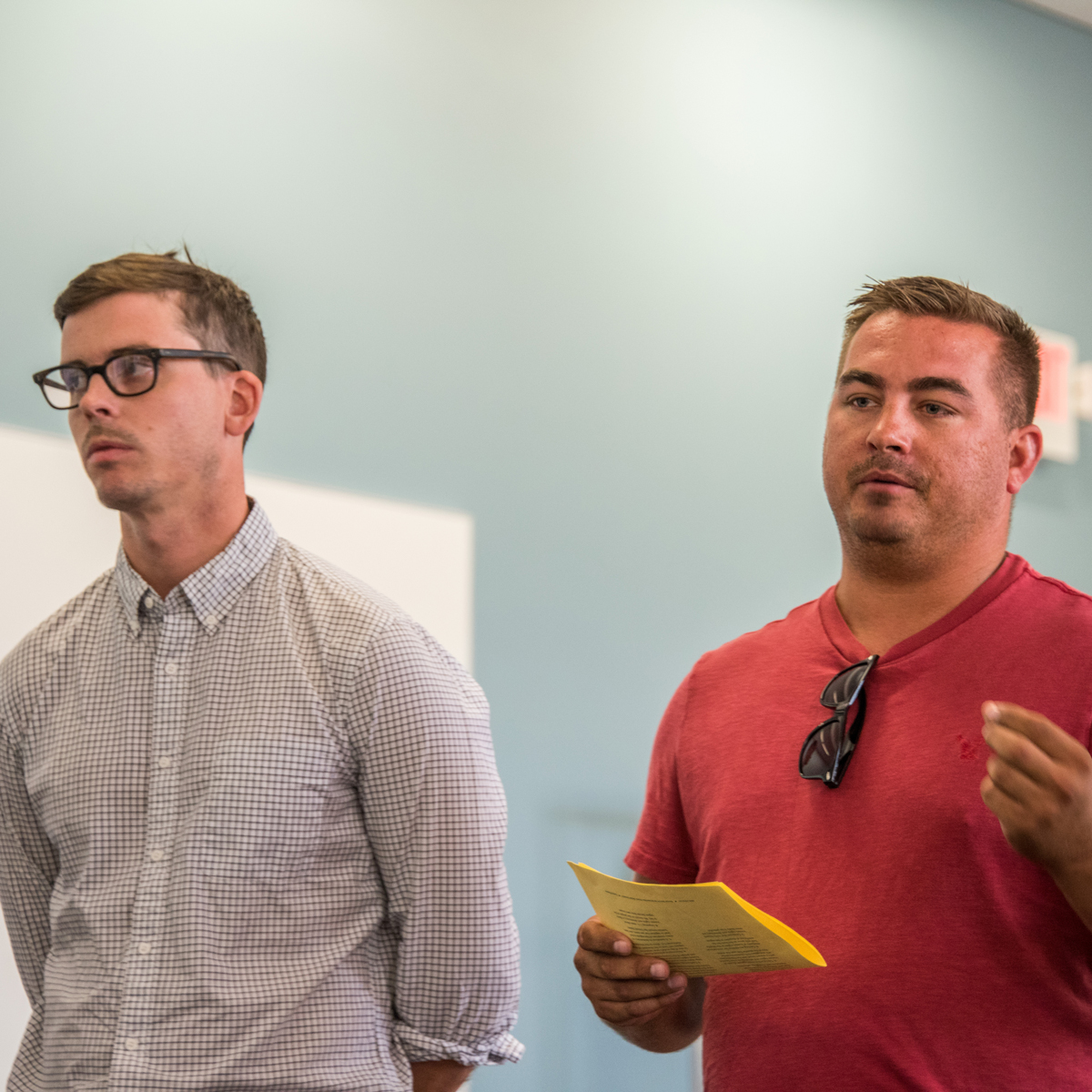 Two men standing. One holds notes and is talking