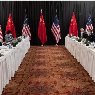 U.S. and China dignitaries seated at two long tables facing each other with several U.S. and Chinese flags in the background.