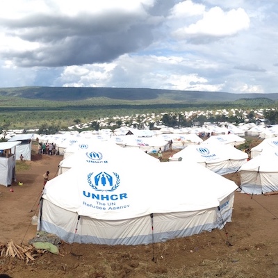 The UNHRC Mahama refugee camp in Rwanda with rows and rows of white tents lined up on dirt and a few people milling about. At one point, the camp housed roughly 48,000 people.