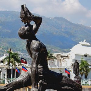 The statue of Le Marron Inconnu in Haiti showing a freed person blowing a conch shell pointed to the sky. The statue is set against a backdrop of buildings, palm trees, and hills.