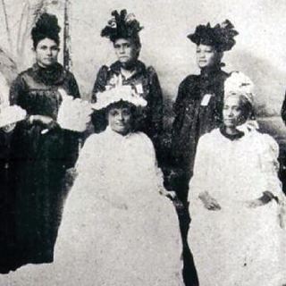 Black and white photo of members of the Hawaiian Patriotic League for Women pose in two rows, one seated and one standing; most wear fancy dresses and ornate hats/headpieces