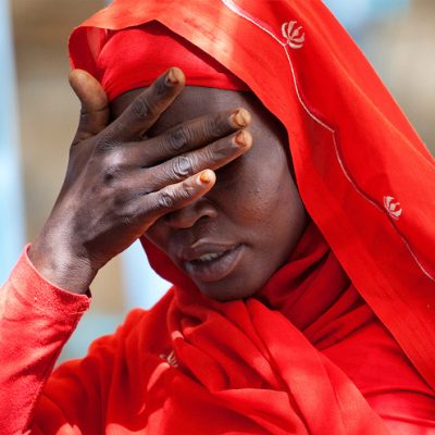 An African woman, dressed in a red dress and head covering, holds her hand over her eyes.