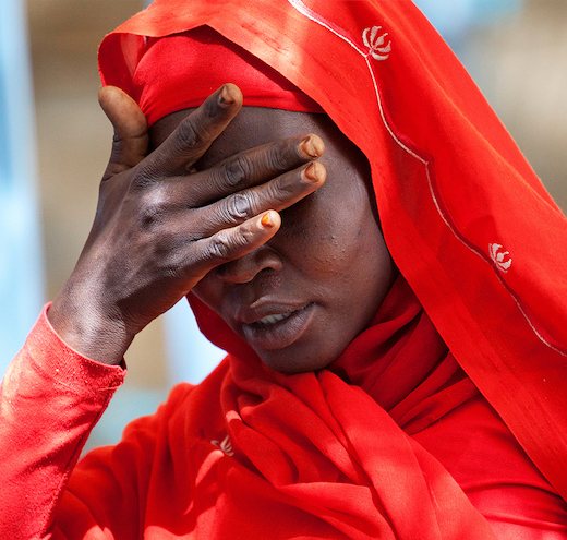 An African woman, dressed in a red dress and head covering, holds her hand over her eyes.