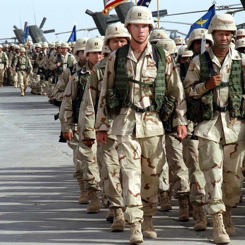 Dozens of U.S. soldiers in formation march along a road in front of large helicopters in Iraq in 2003.
