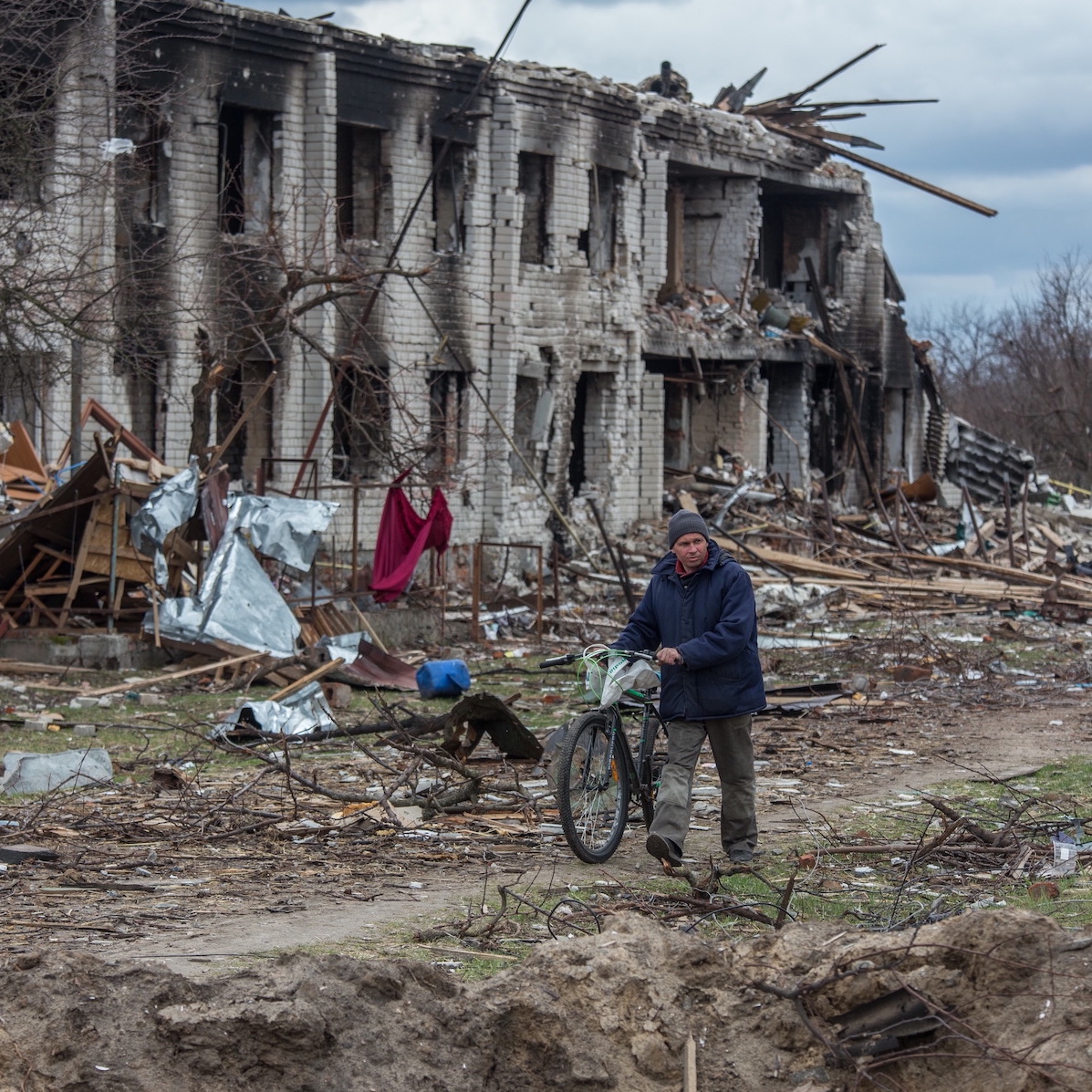 A man in a winter coat and hat pushes his bicycle along a dirt path in front of a bombed out building in Ukraine. Debris and tree branches litter the ground.