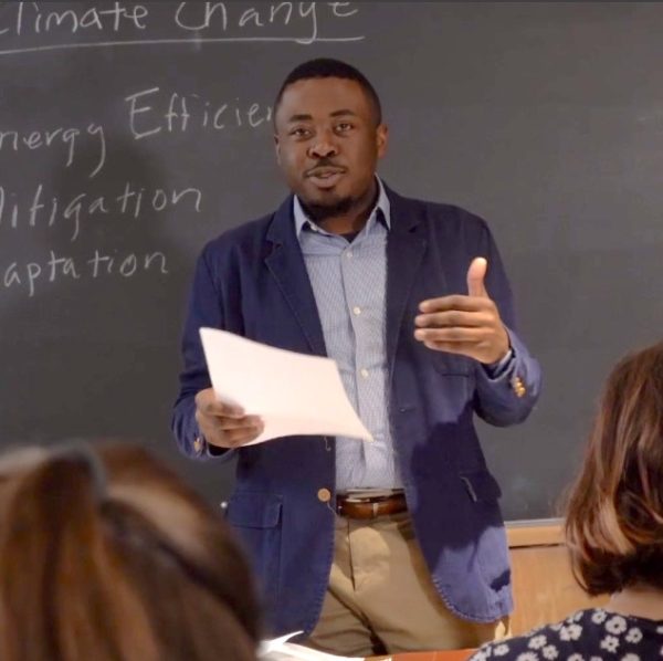 A Black teacher stands at the front of the classroom in front of chalkboard with terms related to climate change written on it.