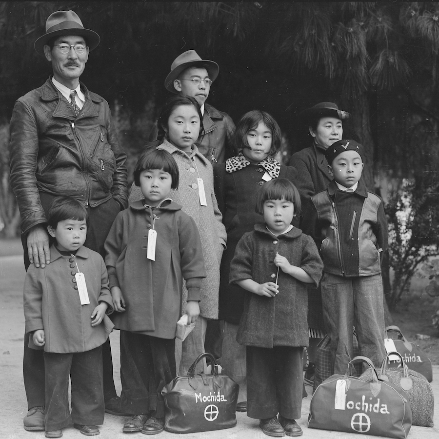 A black and white photo of a Japanese American family of nine standing in a group wearing winter coats with a few small packed bags at their feet marked with the name Mochida. Most of the children have tags attached to their clothing.