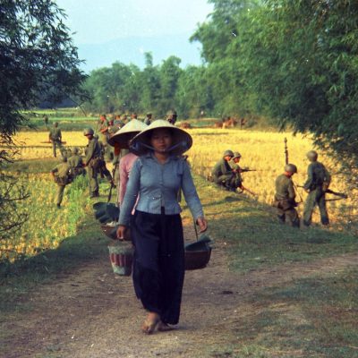 A row of Vietnamese women walk along a path between two fields, heavily patrolled by U.S. Marines in 1965.