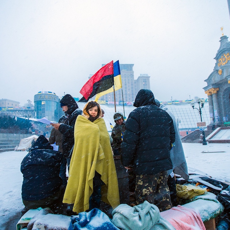A small group of people huddle in a circle in the snow in Russia holding the Ukraine flag and a red and black flag, protesting the war in Ukraine.