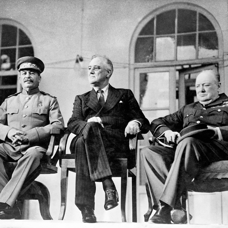 Black and white photo of Stalin, FDR, and Churchill seated in a row during their WWII talks in Tehran in 1943.