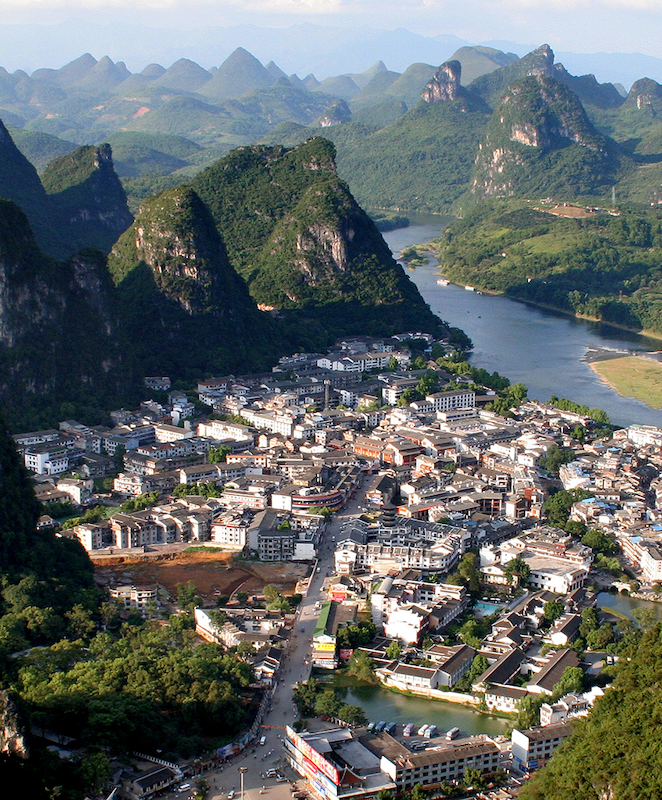 Aerial shot of the town of Yangshuo in China with steep hills surrounding the buildings and a nearby river. Everything looks very lush and green.