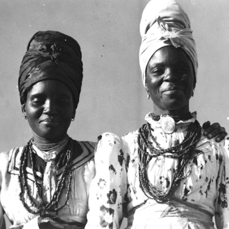 Black and white closeup of two Namibian women in the early 1900s wearing a combination of traditional and German-influenced clothing.