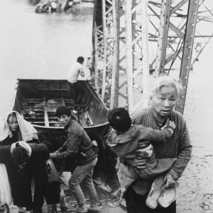 Black and white photo fo Vietnamese civilians fleeing the Battle of Hue in 1968. They are exiting a wooden boat after crossing a river beside a bombed out bridge. The group includes the elderly and small children.