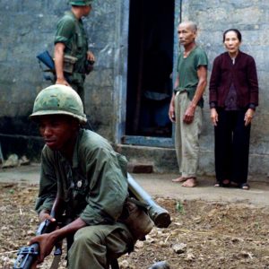 An older Vietnamese stand against the outside wall of their concrete block home while U.S. troops conduct a search of their home in rural South Vietnam in 1968. Two soldiers keep guard over them and the surrounding area.