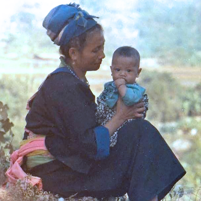A color photo of a Hmong woman sitting on the ground holding a small child on her lap. Laos, 1973.