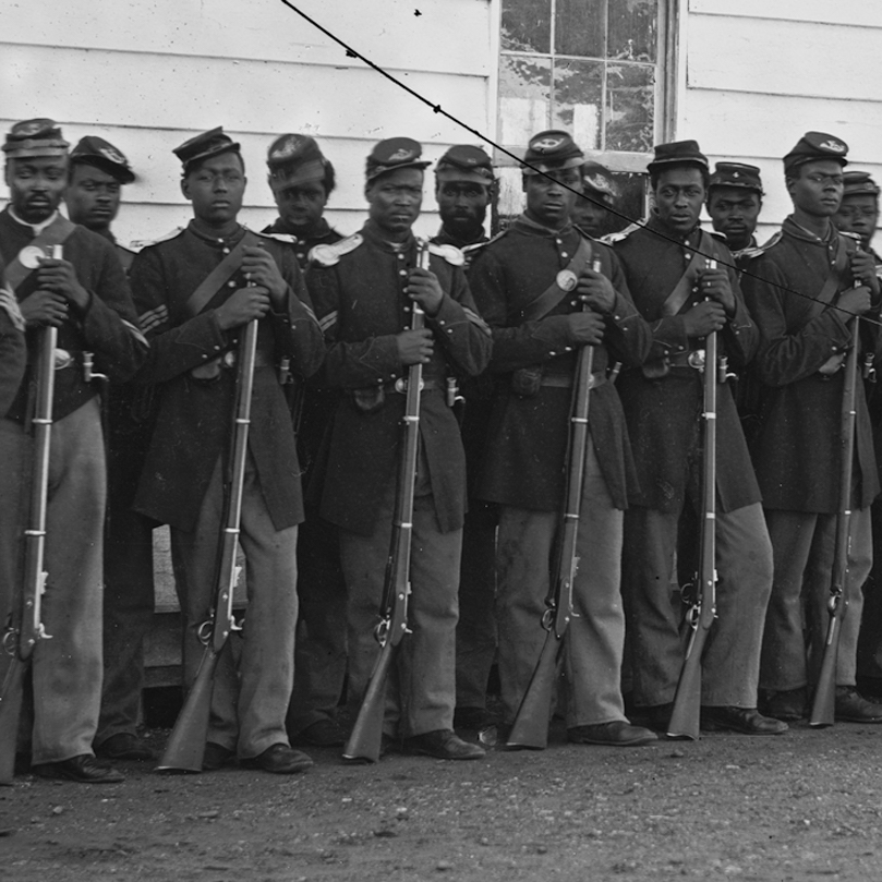 A black and white photo of a row of Black Union soldiers standing in front of a clapboard building with their rifles.